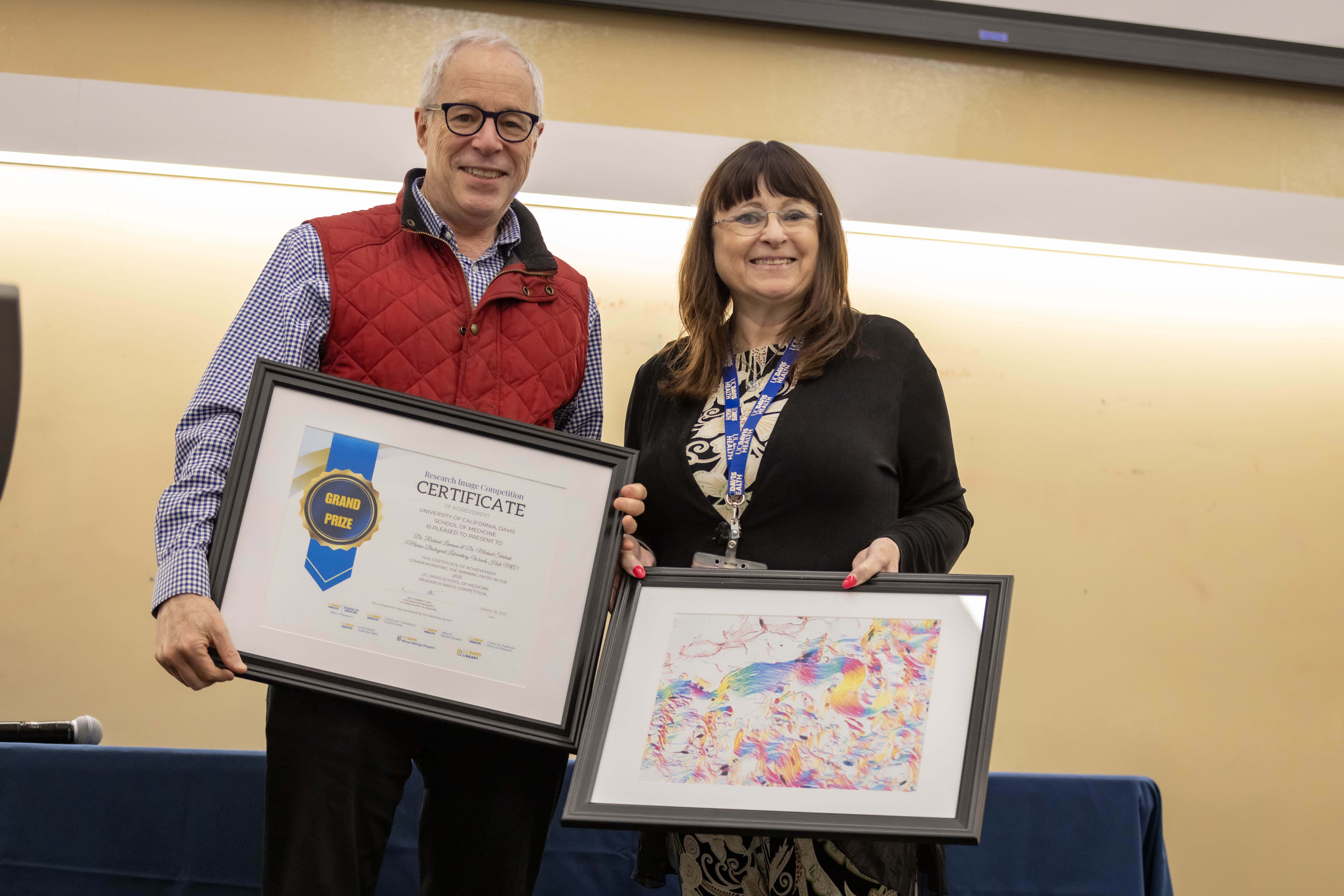 A man in a red vest holding a large framed certificate stands next to a woman in a suit holding a framed microscopy image